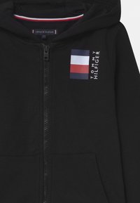 Felpa nera con zip realizzata in morbido tessuto, caratterizzata da un piccolo logo Tommy Hilfiger con un design a bandiera rettangolare nei colori rosso, bianco e blu.