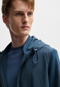 Leichte blaue Zip-Jacke mit Kapuze, mit strukturierten Ärmeln, Zugbandverstellung und einem schlichten blauen Rundhals-T-Shirt darunter.