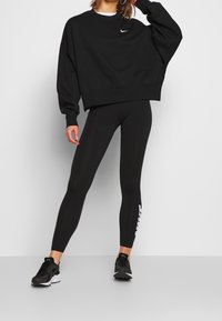 Black oversized sweatshirt with a small white logo, paired with black leggings featuring a white side graphic, and black athletic shoes.