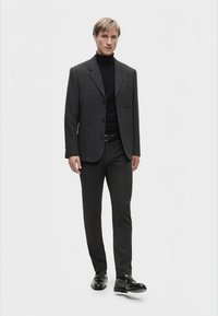 Abito grigio scuro con giacca strutturata e pantaloni abbinati, abbinato a un dolcevita nero e scarpe in pelle nere. Design semplice e sartoriale.