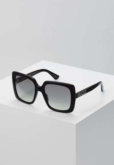 Melni pārāk lieli saulesbrilles ar gradienta pelēkajiem stikliem. Biezas rāmis un sānu rokturi, kas izrotāti ar "Gucci" logotipu, izgreznotu ar rhinestone akmeņiem.