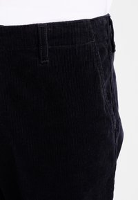 Gros plan sur la taille de pantalon en velours noir et la poche supérieure, montrant la texture du tissu et la boucle de ceinture sur fond blanc.