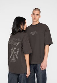 Mørkebrune oversized t-shirts i bomuld med korte ærmer. Foran pryder brandlogoet; bagsiden viser grafik med tekst og dekorative elementer.