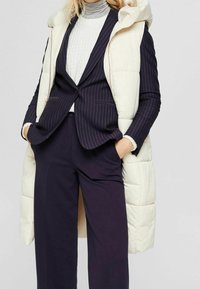 Kobieta ubrana w białą, pikowaną kamizelkę bez rękawów, na granatową blazer w prążki, białą sweter z prążkami i granatowe szerokie spodnie, z rękami w kieszeniach.