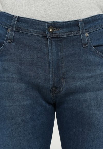 Tamno plave traperice s prednjim kopčanjem, vidljivim remenim petljama i prednjim džepovima, nosi se s svijetlosivim topom.