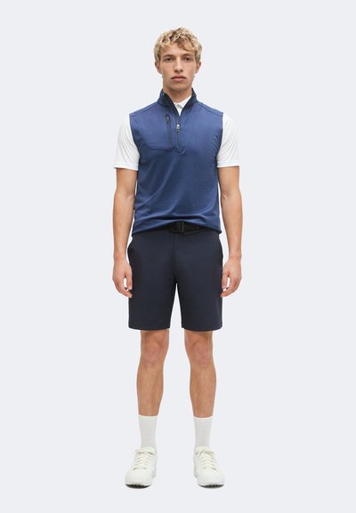 Homme portant une veste sans manches bleue zippée par-dessus une chemise blanche à manches courtes, un short noir, des chaussettes blanches et des baskets blanches, debout devant un fond clair.