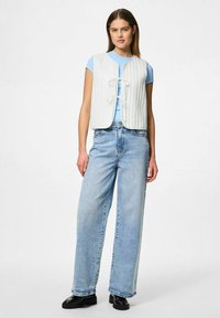 Vitt quiltat väst med framknäppning, bärs över en ljusblå topp, kombinerat med ljusblå vida jeans och svarta platåskor.
