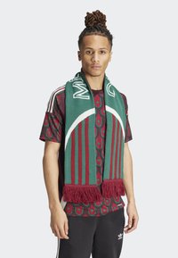 Bufanda de lana verde y burdeos con flecos, que presenta un patrón audaz y acentos de texto. Usada sobre una camisa estampada y pantalones negros.
