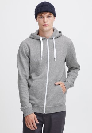 Grijze zip-up hoodie van zacht materiaal met witte trekkoorden, kangoeroezak en geribbelde manchetten, gedragen met een marineblauwe beanie.