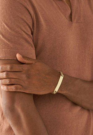 Bracciale in barra d'oro con accenti di catena intrecciata su un polso, esposto su una camicia marrone texturizzata. Superficie liscia e finitura lucida.