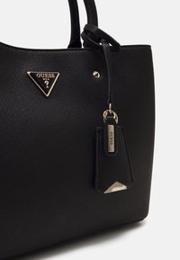 Sac à main Guess en cuir noir texturé avec logo triangulaire doré et étiquette en cuir assortie suspendue à la poignée.