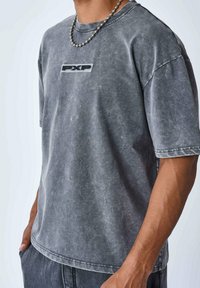 Camiseta de algodón gris, de gran tamaño, con una textura desgastada y un cuello redondo. Presenta un logotipo negro de "PXP" en el frente y mangas cortas.
