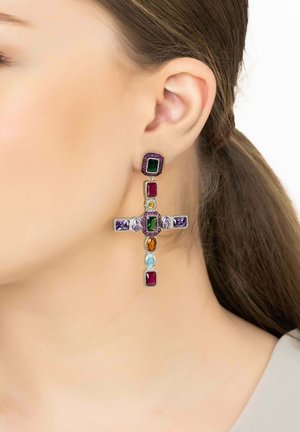 Oreille d'une femme portant une grande boucle d'oreille en forme de croix avec des pierres précieuses multicolores, comprenant du vert, violet, rouge, orange et bleu.