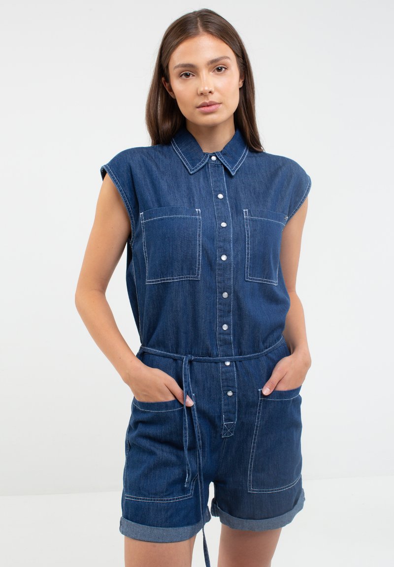 Džinsinis kombinezonas su trumpomis rankovėmis, priekyje su mygtukais, dviem krūtinės kišenėmis ir suveržtu liemeniu. Tamsiai mėlynos spalvos su matomomis siūlėmis ir apvadu.