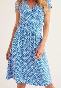 Blå omlottklänning med ett vitt vågmönster, korta ärmar, en figursydd överdel och en utsvängd kjol som slutar ovanför knäet. Tillverkad av bomull.