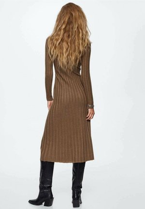 Femme aux longs cheveux ondulés portant une robe midi en maille côtelée marron et des bottes noires montant jusqu'aux genoux, debout et tournée de dos.