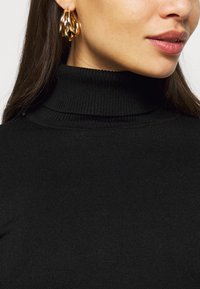 Must mustriba turtleneck 'sviiter musta värviga, millel on kõrge kaelus ja sile tekstuur. Kullast rõngas kõrvarõngad on nähtavad, tuues sisse metallset detaili.