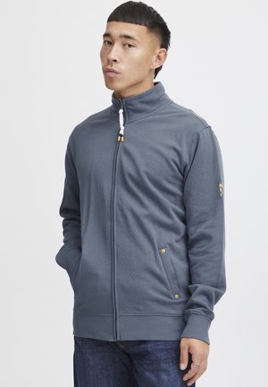 Grå zip-up hettegenser med høy krage, to sidelommer og detaljer i gulltone; teksturert stoff og ribbestrikkede mansjetter gir et glitrende utseende.