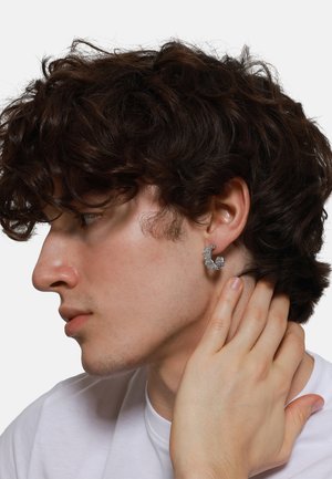 Boucle d'oreille en métal argenté avec un design abstrait et ondulé, portée à l'oreille gauche. La personne a les cheveux bruns bouclés et une expression neutre.