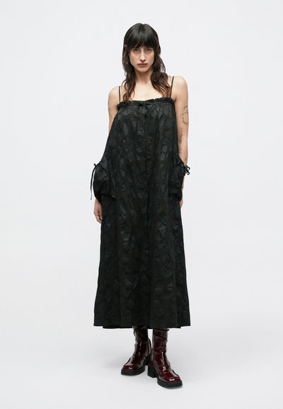 Μαύρο μακρύ φόρεμα με υφή υφάσματος, ρυθμιζόμενα κορδόνια στους ώμους και τα μανίκια, συνδυασμένο με γυαλιστερές μπορντό μπότες αστραγάλου.