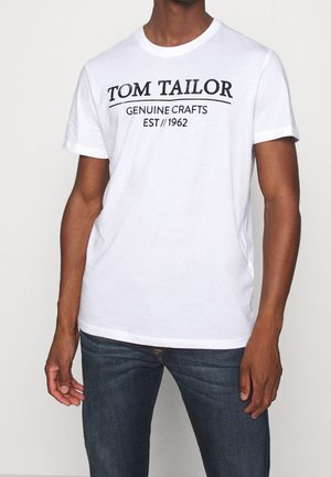 Mężczyzna w białej koszulce z krótkim rękawem z czarnym napisem "TOM TAILOR GENUINE CRAFTS EST 1962" i ciemnoniebieskich dżinsach, stojący na jednolitym tle.