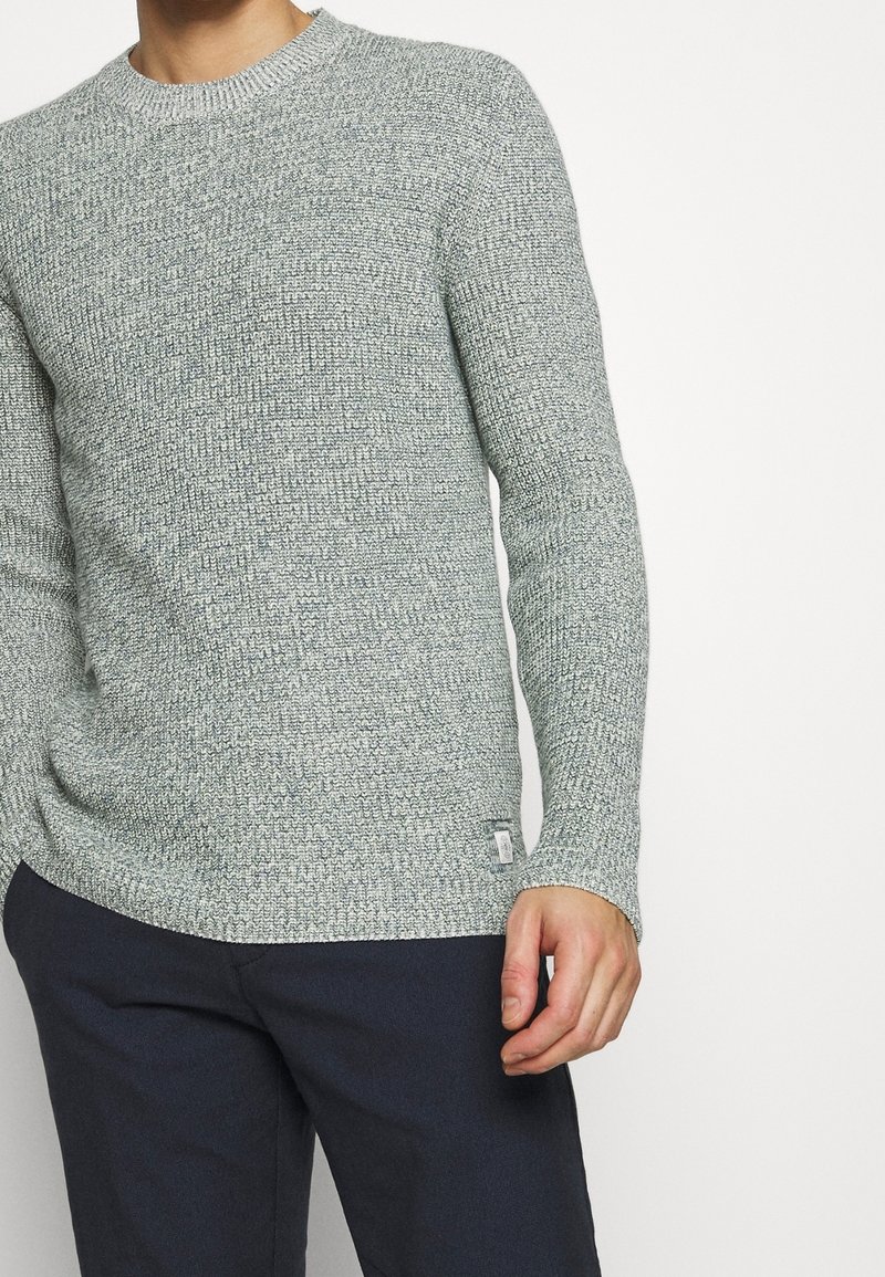 Pull en tricot gris clair avec un motif texturé, col rond et manches longues. Porté sur un pantalon sombre, avec une petite étiquette à l'ourlet.