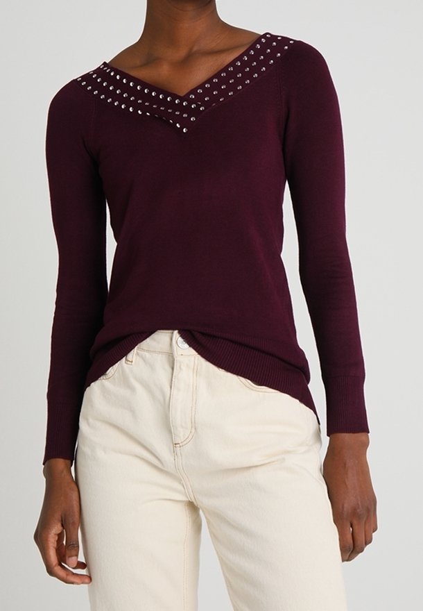 Burgundy v-neck sweater with decorative silver studs along the neckline. Long sleeves and ribbed hem, paired with light-colored pants.