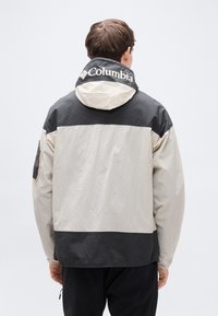 Ε λαφρύ μπουφάν σε μπεζ και μαύρο, με κουκούλα και ελαστικά μανίκια. Διαθέτει λογότυπο στο γιακά και λεπτομέρεια τσέπης στο μανίκι.