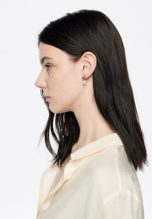 Pendientes de studs en tono dorado con un diseño geométrico texturizado y una perla blanca suspendida, combinados con una blusa ligera y transparente.