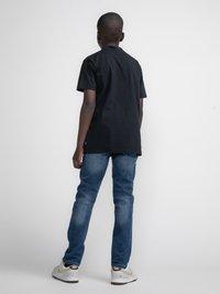 Tricou din bumbac negru, croială lejeră, purtat cu blugi albaștri. Pantofii sunt albi cu accente aurii. Fundal simplu, concentrat pe îmbrăcăminte.