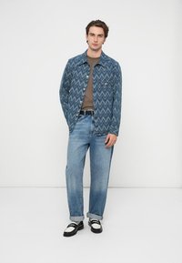 Denimtakki siksak-kuviolla, keskisininen; ruskea paita; vaalean siniset farkut; mustavalkoiset kengät; etunäkymä, seisova miesmalli.