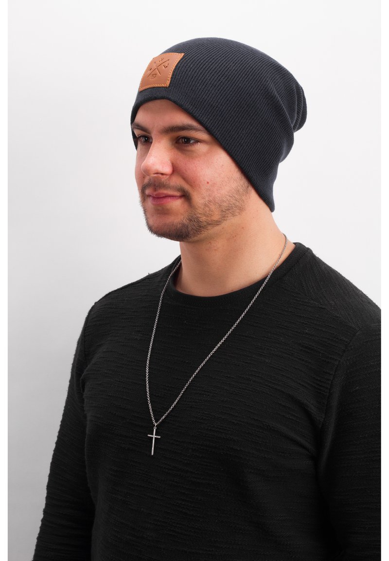 Dark blue ribbed knit beanie with a tan leather patch at the front. The wearer is in a textured black sweater and a silver chain necklace.