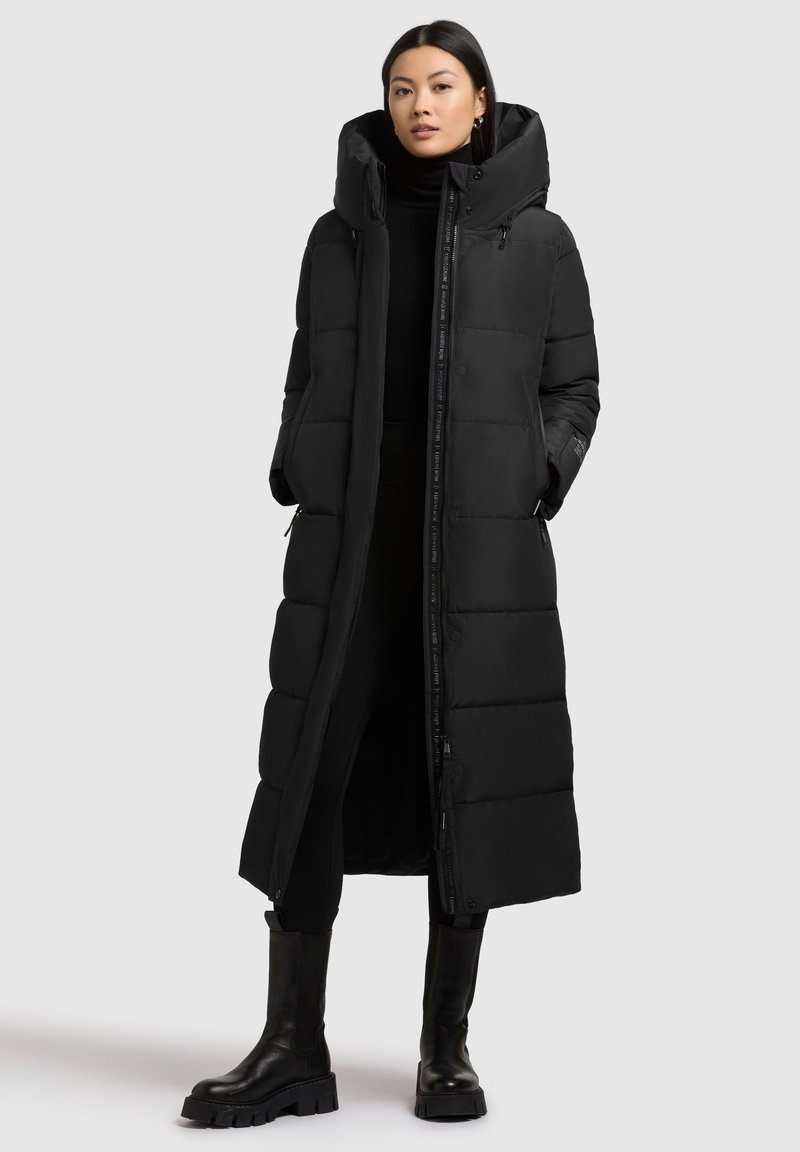 Sort lang puffer jakke med høj krave, lynlås lukning og trykknapper. Har quiltet tekstur og sidelommer. Sammen med sorte støvler.