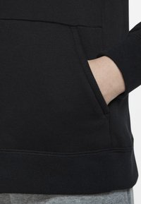 Črna majica s kapuco z kengurujskim žepom, rebrastimi manšetami in teksturirano tkanino. Rob je rahlo daljši zadaj in je v paru s sivimi trenirkami.