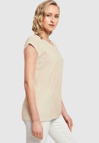 Beige V-hals T-skjorte med korte ermer og en jevn tekstur. Merkelig detalj: liten trykt logo foran. Kombinert med hvite bukser.