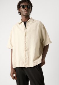 Lichtbeige kortebloemige overhemd met een spread collar en zes knopen. Vlotte textuur, gedragen over een zwarte basislaag, met een losse pasvorm.