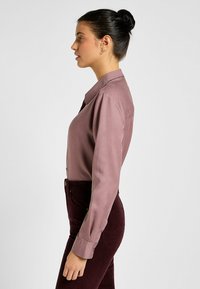 Koszula w kolorze mauve z długimi rękawami i kołnierzykiem zapinanym na guziki, zestawiona z ciemno-burgundowymi spodniami. Materiał wydaje się gładki z subtelnym połyskiem.