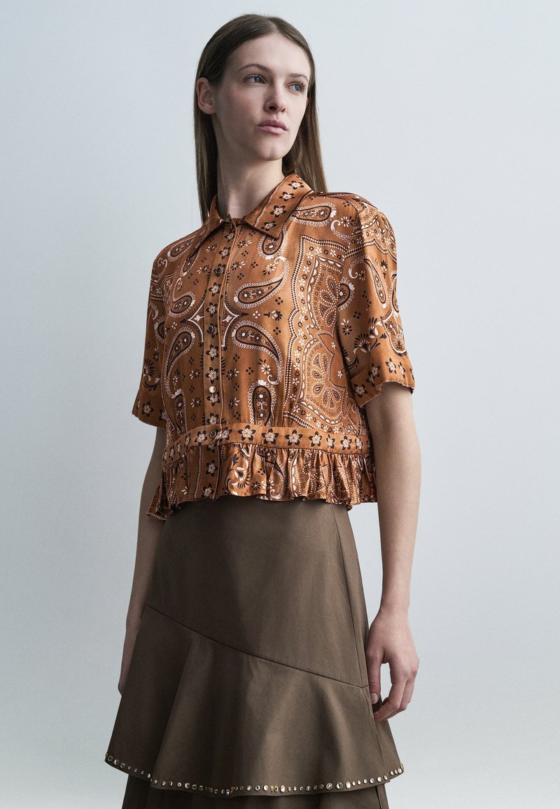 Νεαρή γυναίκα που φοράει μπλούζα με κοντά μανίκια σε πορτοκαλί μοτίβο πεϊσλί και φούστα καφέ ψηλόμεση με διακόσμηση από μεταλλικά καρφιά.