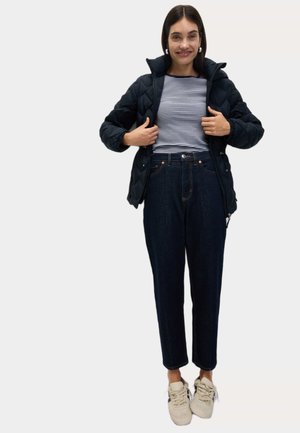 Sort quiltet jakke over en marineblå og hvid stribet skjorte, parret med højtaljede mørke jeans og beige sneakers.