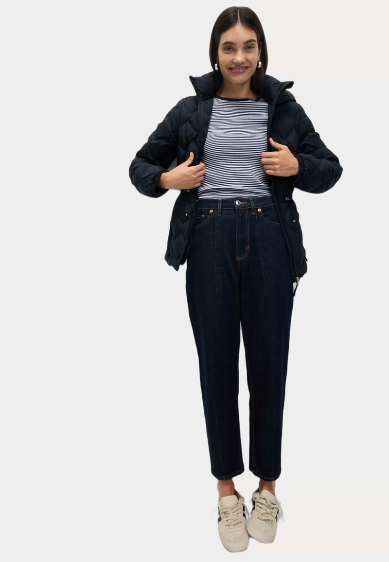 Černá prošívaná bunda přes námořnickou a bílou pruhovanou košili, v kombinaci s vysokým pasem tmavými džínami a béžovými teniskami.