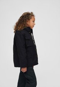 Veste en toile noire avec une fermeture éclair et des boutons, dotée de deux poches avant et d'un col haute. L'enfant porte un pantalon foncé.
