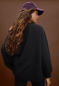 Felpa nera oversize con spalle scese, caratterizzata da orlo e polsini a costine. Abbinata a un cappellino da baseball viola.