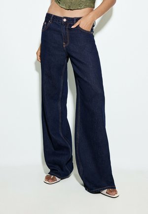 Femme portant un jean bleu foncé à jambes larges, un top court vert et des sandales blanches à talons ouverts, debout sur un fond blanc.
