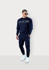 Navy Sweatshirt mit "ESTEEM"-Logo, kombiniert mit passenden Jogginghosen. Hergestellt aus weichem Stoff, mit gerippten Bündchen und Kordelzugbund. Weiße Sneakers.