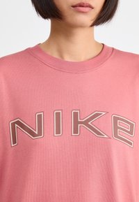 Růžové bavlněné tričko s výrazným béžovým logem "NIKE" s konturovým detailem a designem s kulatým výstřihem. Hladká textura s krátkými rukávy.