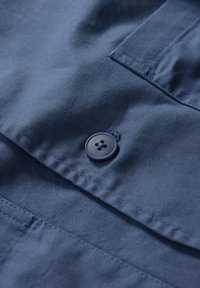 Navy blue cotton fabric with a smooth texture, featuring a circular, four-hole button and visible stitching along the edges and pocket.