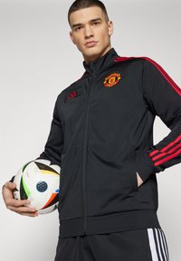 Sort atletisk jakke med røde striber, Manchester United-logo på brystet, båret af en person der holder en multicoloret fodbold.