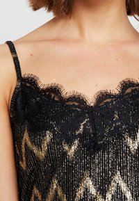 Černý top s tenkými ramínky a metalickými zlatými detaily. Má krajkový lem a dekorativní třásně podél výstřihu. Vertikálně pruhovaná textura.