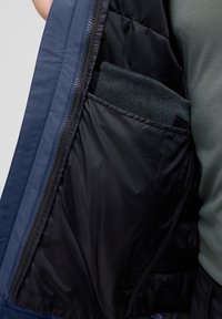 Veste isolée bleu marine avec une poche en tricot gris visible à l'intérieur. L'intérieur présente une doublure noire lisse et une fermeture éclair le long du bord.
