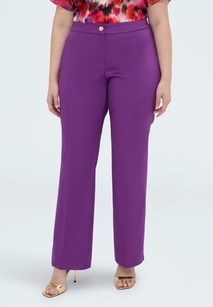 Femme portant un pantalon violet taille haute à jambes droites, associé à des sandales à talons ouvertes dorées et un top fleuri coloré.
